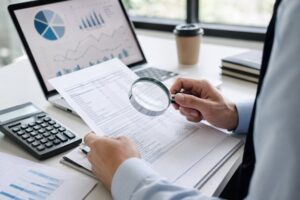 Businessman examining financial reports with a magnifying glass to uncover hidden costs before purchasing products or services.