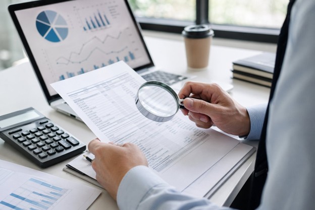 Businessman examining financial reports with a magnifying glass to uncover hidden costs before purchasing products or services.